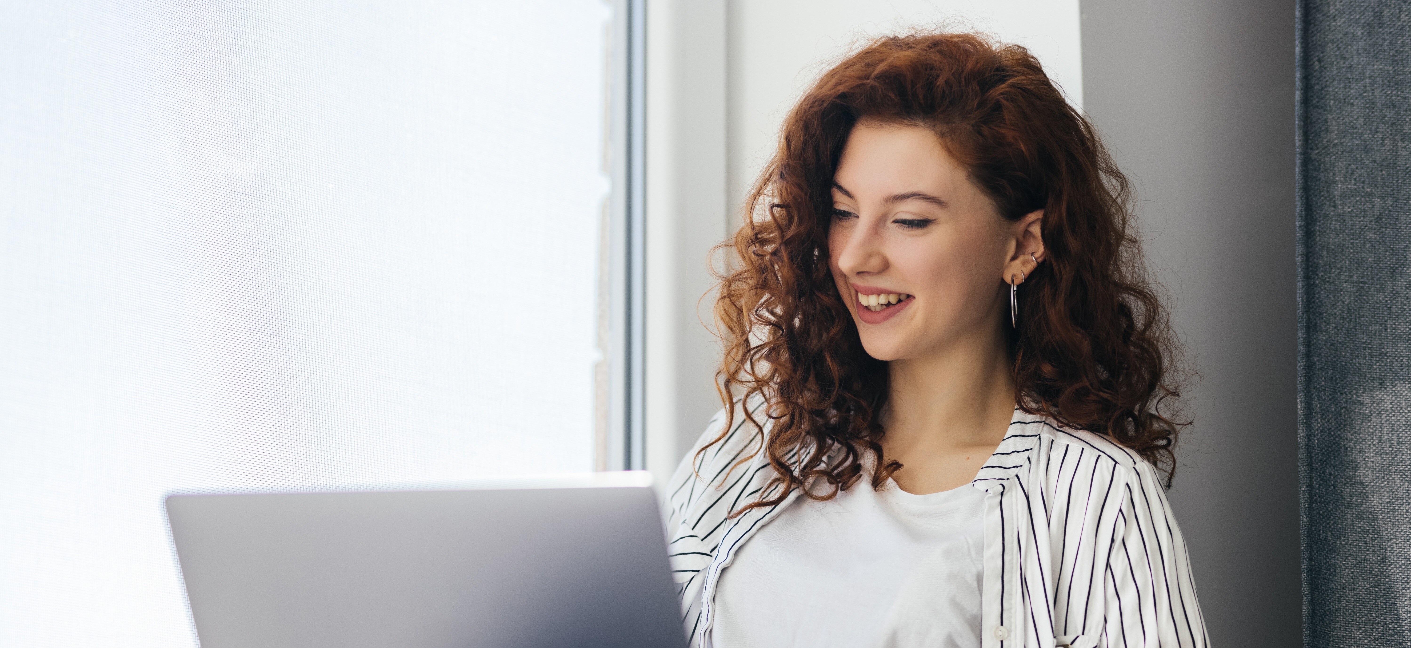 beautiful-positive-woman-using-laptop-sitting-windowsill-city-apartment-young-redhaired-woman-working-home-freelance-concept-1